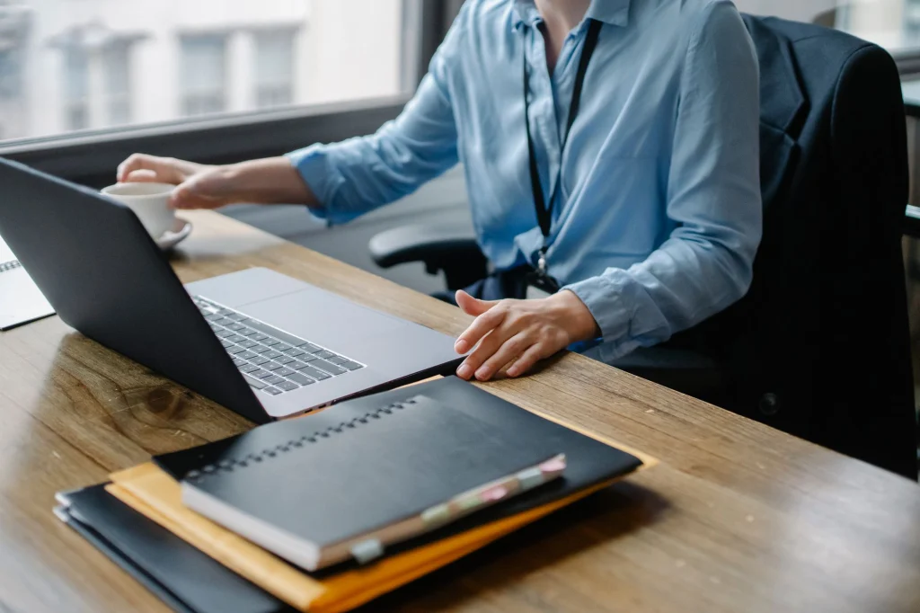 Persoană care lucrează la laptop, cu dosare cu acte pe birou