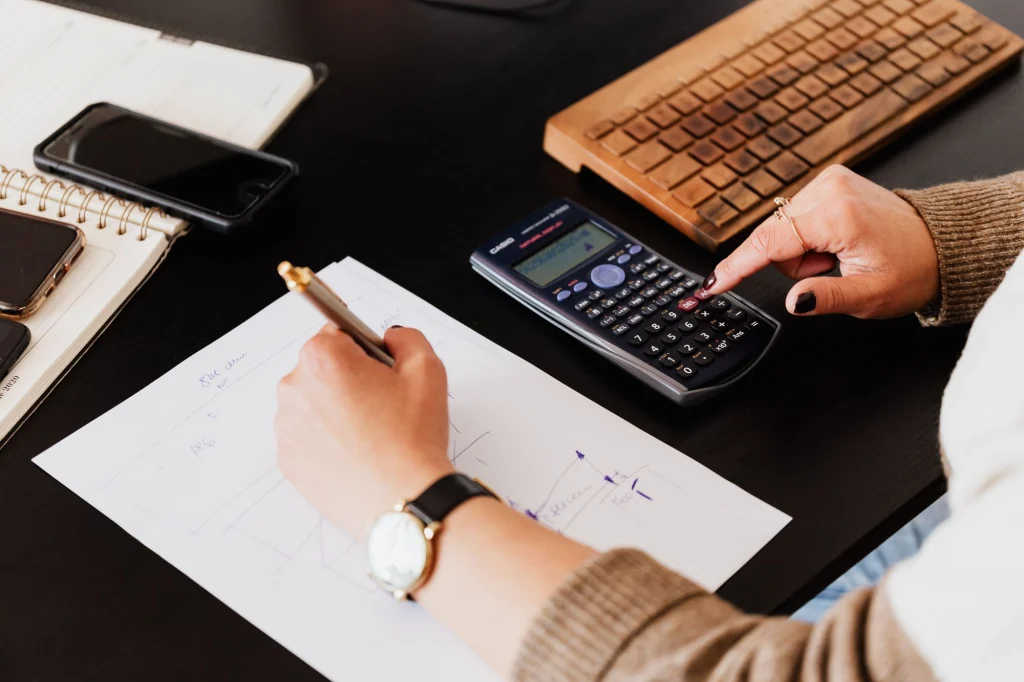 Mâini care notează calcule lângă un calculator, pe un birou